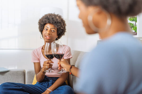Two twin sisters toast wine glasses as they sit on a couch indoors.