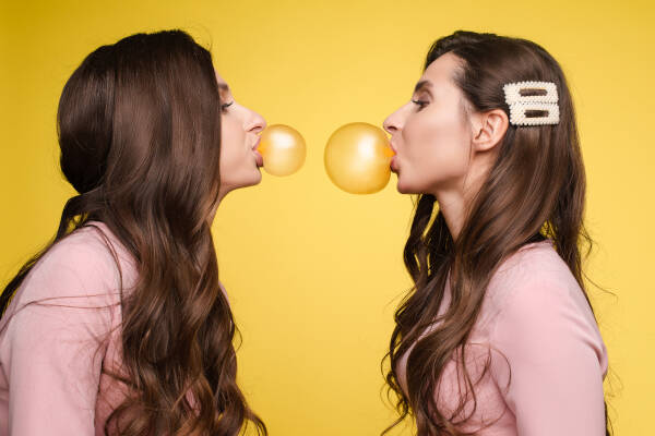 Two twins facing each other, blowing bubbles with bubblegum against a yellow backdrop.