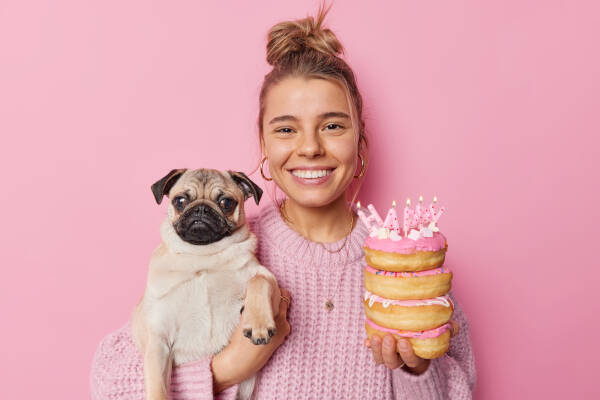 Woman holds a pug dog and doughnuts with candles, smiling against a pink backdrop.