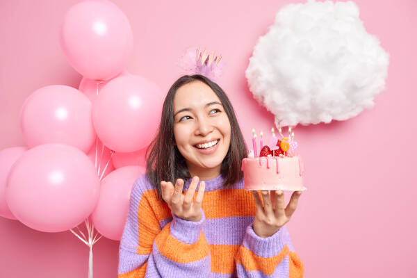 Woman smiles as she holds a small pink cake in her hands against a pink background, next to pink balloons.