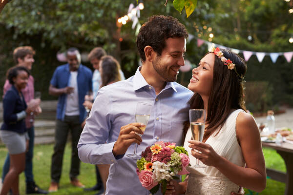 Young couple celebrating their wedding with a backyard party, the man and woman looking into each others eyes as they toast with glasses of white wine.