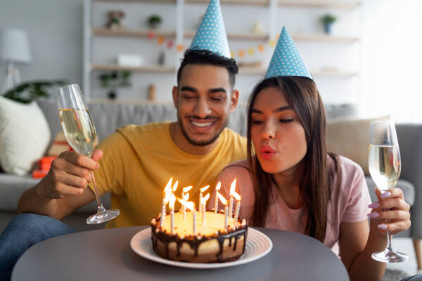 Young couple in party hats at home, girlfriend blows out candles on cake while they both hold champagne glasses.