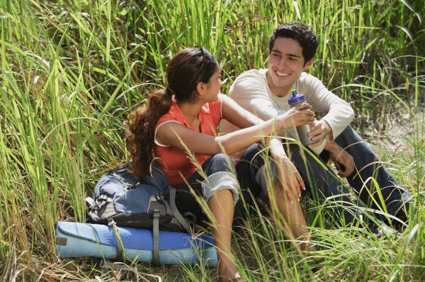 Young couple sitting together with camping equipment, taking a break and smiling in the long grass.