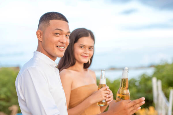 Young couple smile and enjoy a beer as they pose for a photo outside at a party.