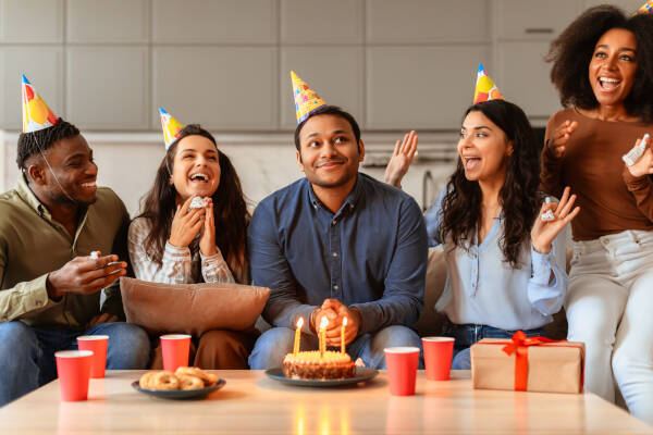 Young friends sat in front of a table of birthday cake, a gift, drinks and snacks, express excitement and joy while celebrating a birthday together at home.