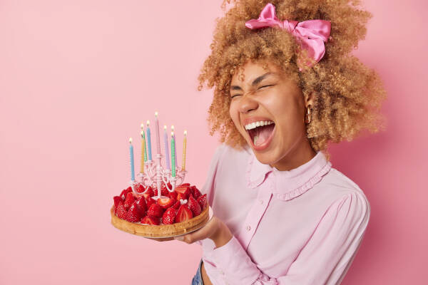 Young woman holds a strawberry cake with candles and exclaims loudly against a pink background.