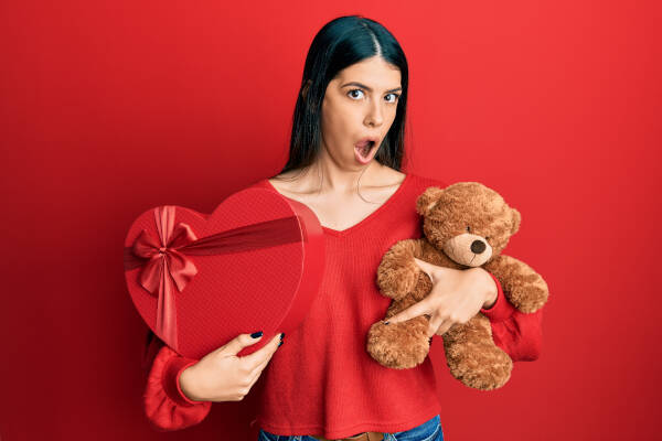 Young woman with a shocked expression, holding a heart-shaped gift box and a teddy bear against a red backdrop.