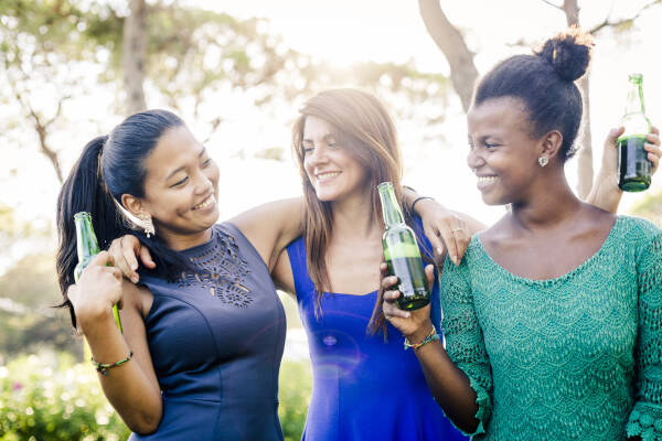 Young women with arms around each other, drinking beers and smiling together.