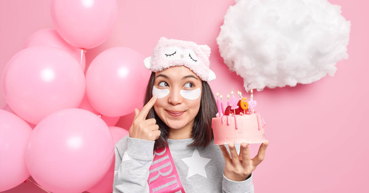 Young woman with beauty patches under eyes holds a cake and wears a birthday sash against a pink backdrop with pink balloons.