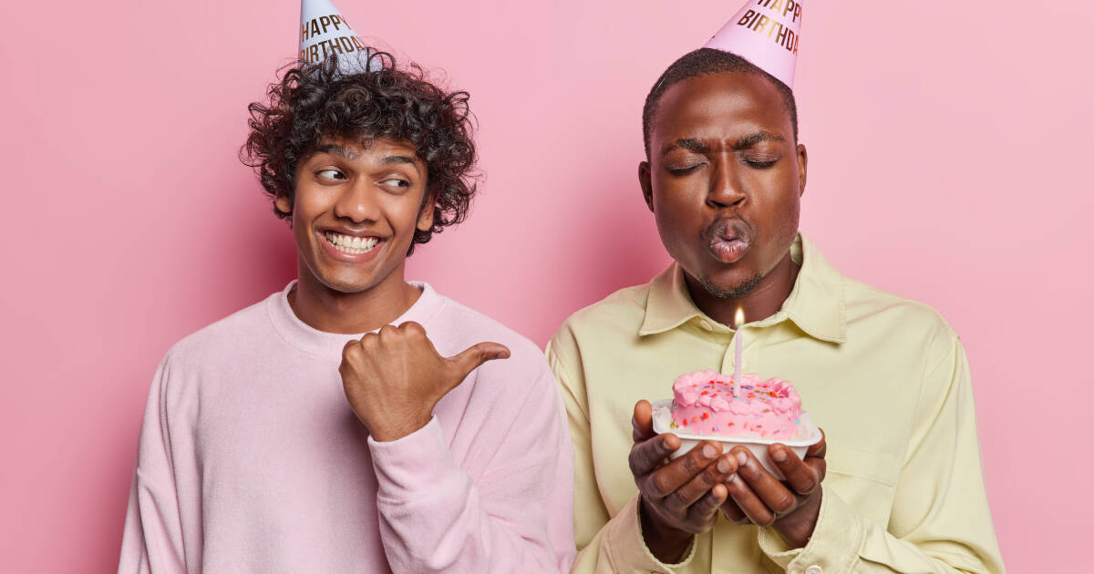 Two young males in party hats stand against a pink background, one blowing out a candle while the other points to him.