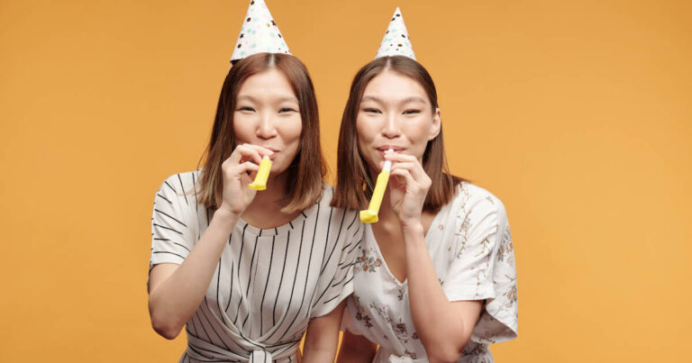 Happy, Young Female Twins wearing party hats and blowing party horns against an orange background.