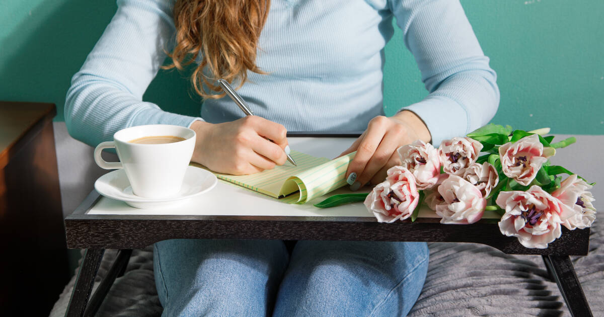 A woman in a light blue shirt writes in a notebook on a tray table with a cup of coffee and a bouquet of pink flowers.