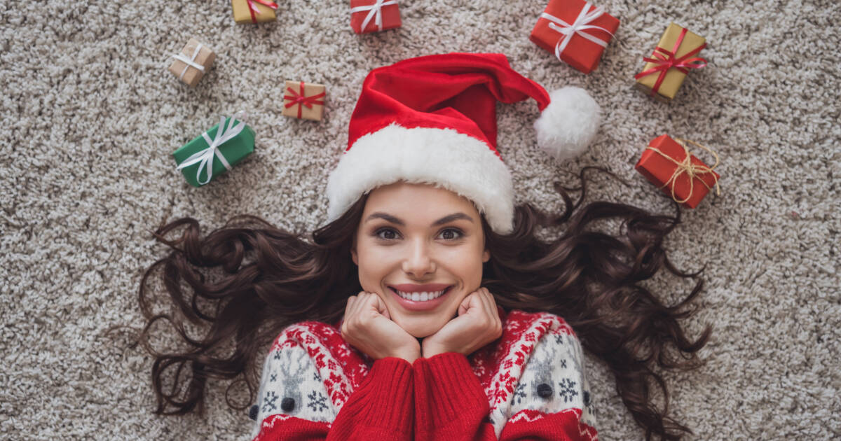 Woman in a Santa hat lying on a carpet, surrounded by small wrapped Christmas gifts, smiling with hands under her chin in a festive sweater.