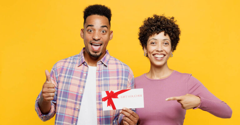 A cheerful couple holding a gift voucher against a bright yellow background. The man shows a thumbs-up, while the woman smiles and points at the voucher.