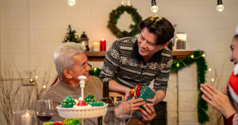 A young man presents a wrapped gift to an elderly man at a festive dinner table, surrounded by holiday decorations and warm lighting, both are smiling.