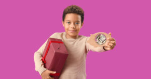 Young boy smiling whilst holding a red gift box under his right arm and a piggy bank in is left hand, set against a pink background.
