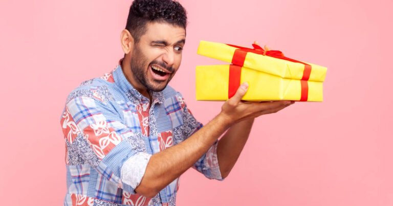 Man squinting with one eye open, looking into a partially opened yellow gift box with a red ribbon, set against a pink backdrop.