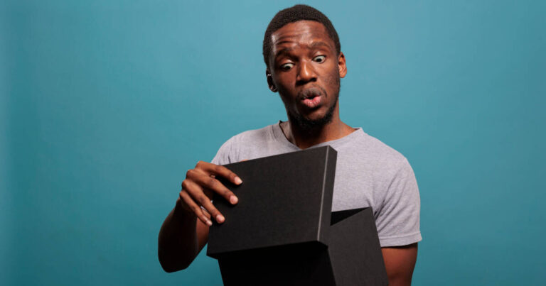 Man peers into a black gift box with a confused expression against a blue background.