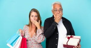 Man and woman hold their hands up to their mouths in shock whilst holding gifts in front of a blue background.