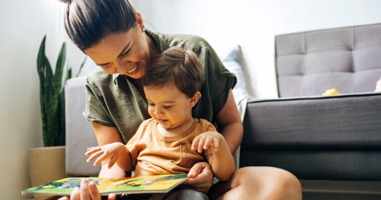 A mother sits on the floor with her young child, both smiling and engaged as they read a colorful picture book together in a cozy room.