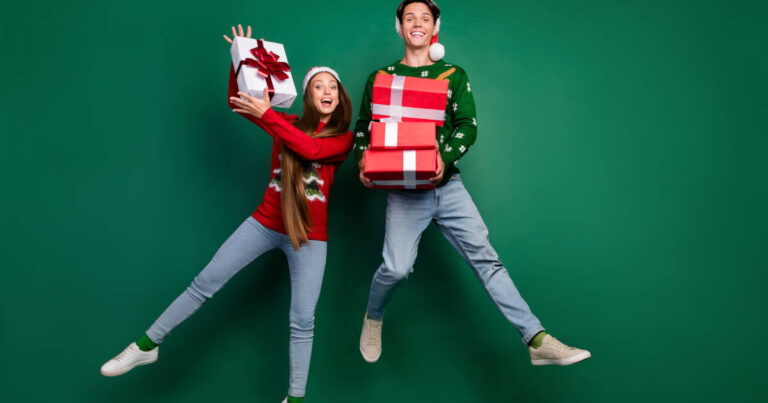 A joyful couple wearing festive Christmas sweaters and Santa hats, enthusiastically jumping against a green background while holding wrapped holiday gifts, ready to celebrate together.