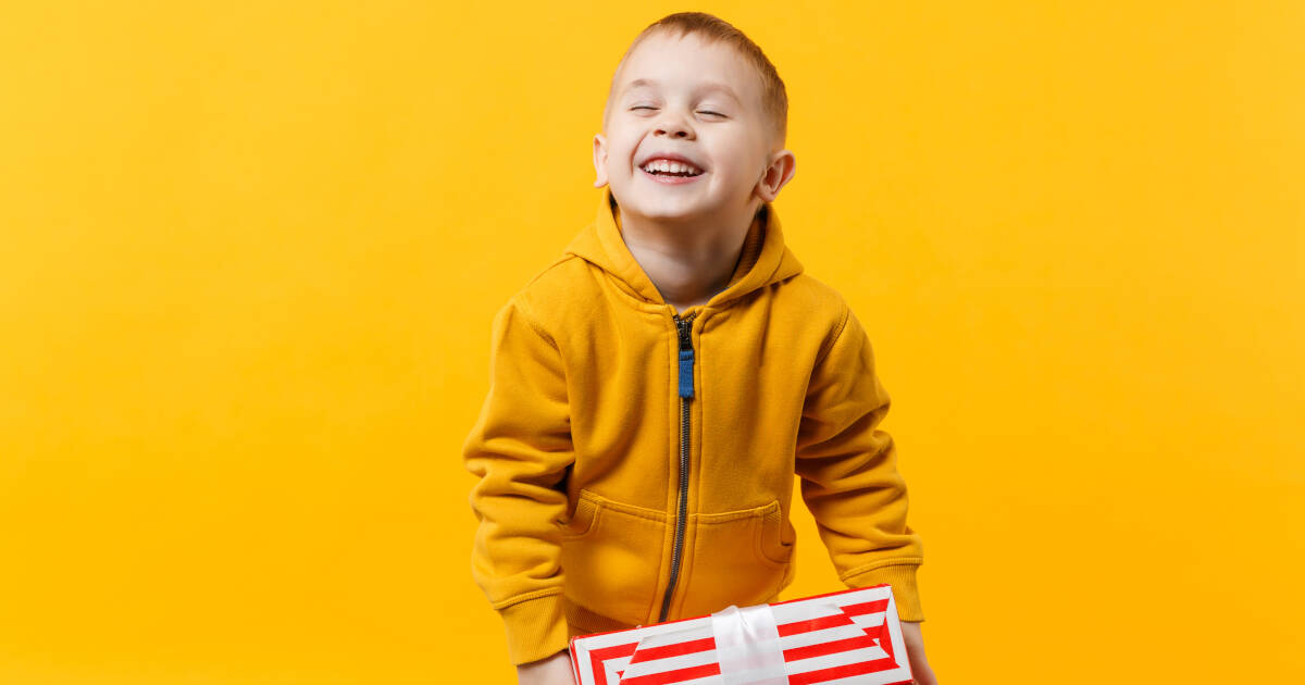 Young boy, wearing a yellow zipped jumper, smiling widely holding a red and silver striped present, set against a yellow backdrop.