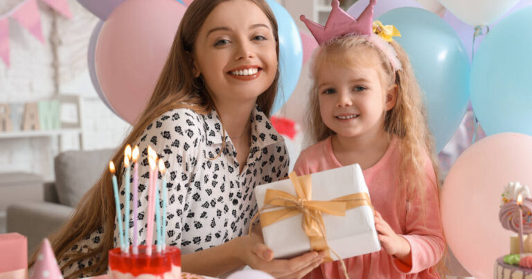 Little girl wearing a pink crown smiling whilst holding a gift with her Mother, standing in front of a candle lit cake.