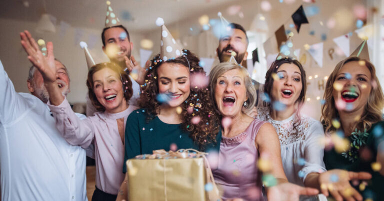 A joyful group of people celebrating at a party wearing party hats and smiling as they throw colorful confetti into the air, a woman in the center holds a gift box wrapped in gold paper.