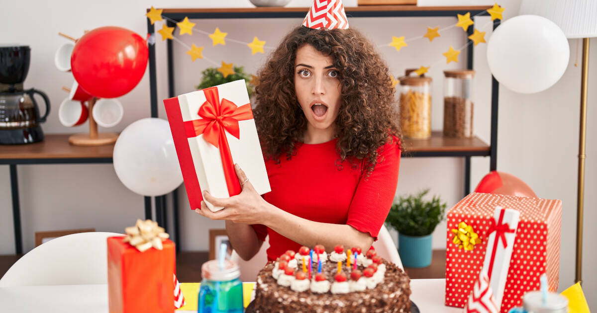 A woman holds up a gift box with a shocked look on her face, with more gifts and a cake on the table at home.