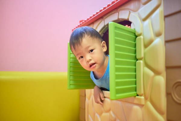 2-year-old toddler boy plays peek-a-boo from the window of a plastic toy house.