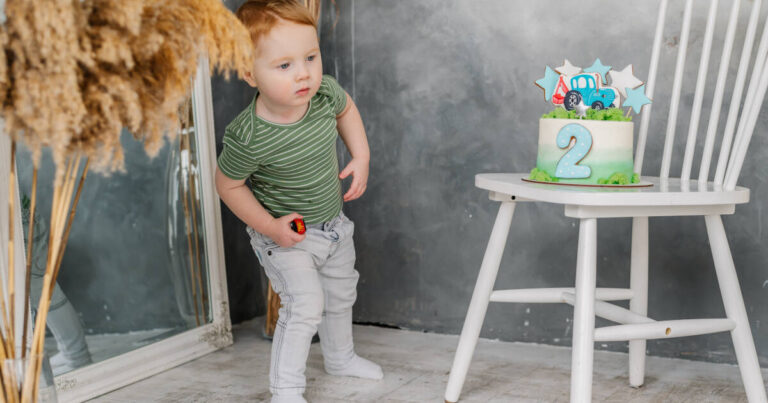 Surprised little boy gazes at trendy 2nd birthday cake on a chair at home.