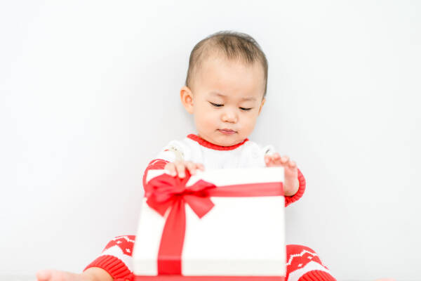 A baby boy smiles as he opens a white gift box with a red ribbon against a white background.