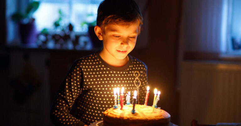 A boy in a dimly lit room wearing a dark shirt with stars, looking at a lit birthday cake with multiple candles.
