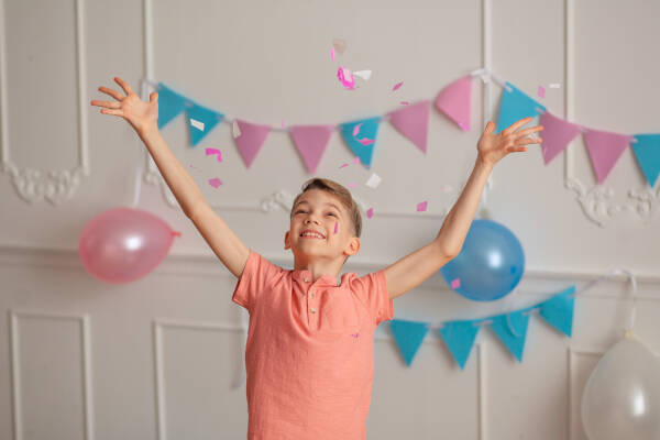 A boy in a pink shirt throwing confetti in the air, celebrating in a room decorated with blue and pink bunting and balloons.