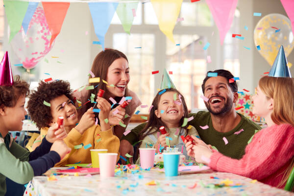 A cheerful group of children and an adult at a colorful indoor birthday party, with confetti falling and party decorations in the background.