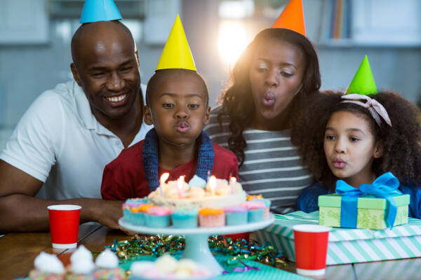 A family celebrating a child's birthday. The child is blowing out candles on a birthday cake, with both parents and a sibling wearing party hats and watching.