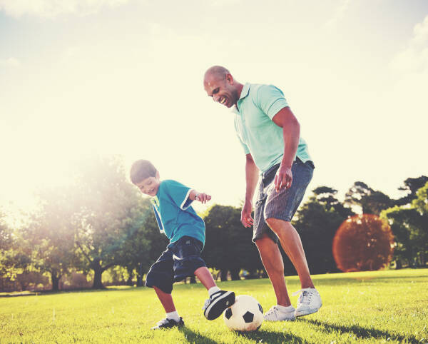 A father and son playing soccer on a sunny day in a park, both smiling and enjoying their time together.