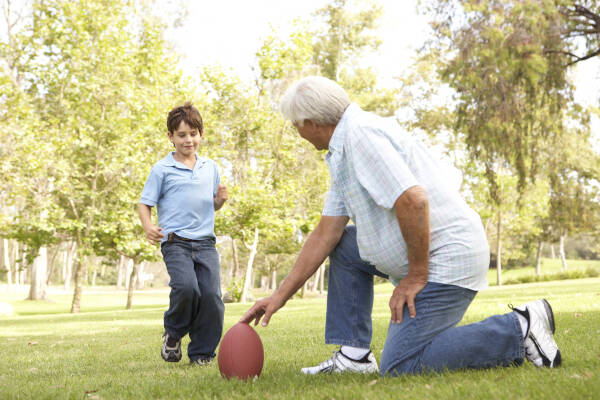 A grandfather and grandson playing with a football in a park, with the grandfather kneeling and the grandson running towards him.