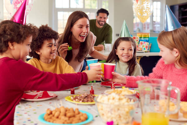 A group of children and an adult at a colorful indoor birthday party, raising cups in a cheerful toast.