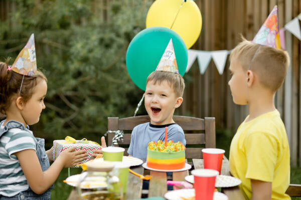 A group of children at an outdoor birthday party. A girl is handing a wrapped gift to a boy, who looks excited.