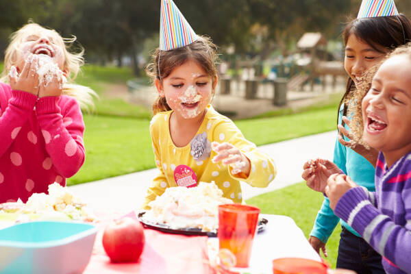 A group of children at an outdoor birthday party. One girl is wearing a party hat and has cake on her face, while others are laughing and enjoying the cake.