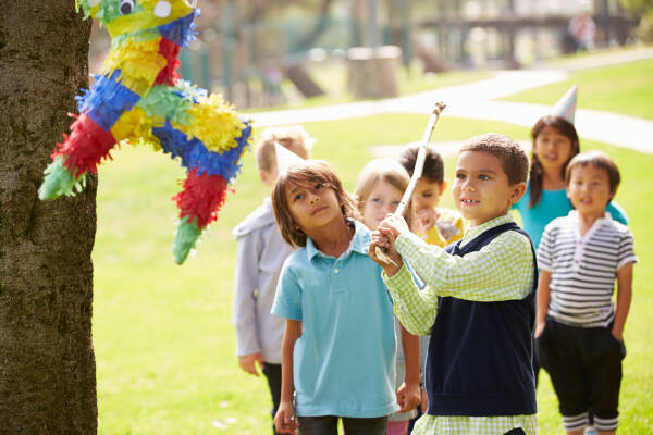 A group of children watching as a boy swings a stick to hit a colorful piñata at an outdoor birthday party.