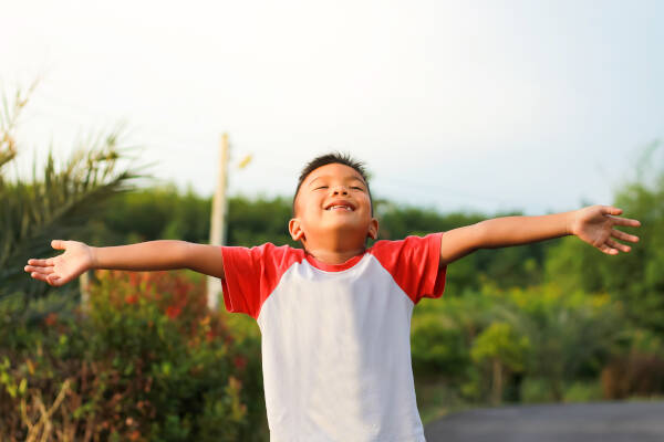 A happy young boy with his arms outstretched, standing outside on a sunny day, looking joyful.