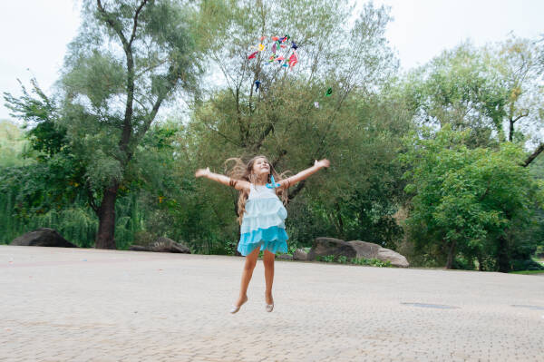 A joyful young girl gleefully jumps and tosses popped balloons into the air outdoors on a sunny summer day.