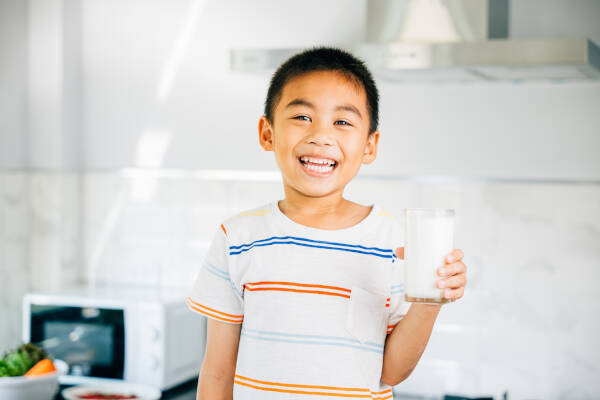 A smiling young boy holding a glass of milk in a bright kitchen, wearing a striped shirt.