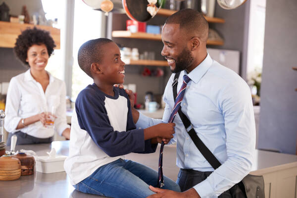 A smiling young boy sitting on a kitchen counter, playfully tugging on his father's tie while his mother looks on, smiling in the background.