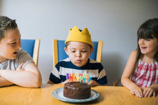 A young boy wearing a yellow crown, sitting at a table, blowing out candles on a birthday cake, with two children sitting beside him.