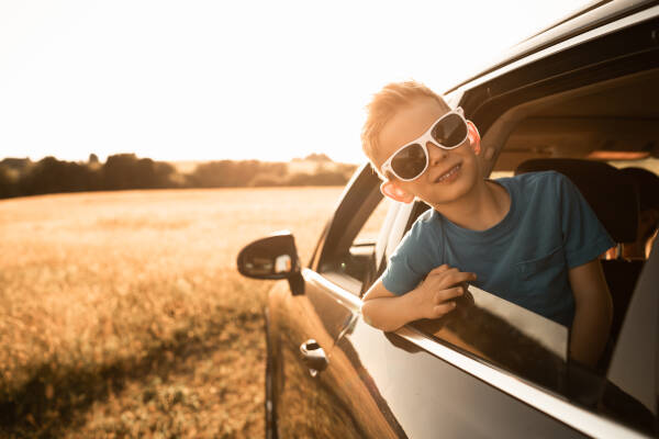 A young boy wearing sunglasses, leaning out of a car window, smiling, with a sunny field in the background.