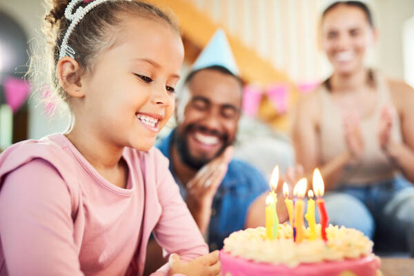 A young girl in a pink shirt smiling at her birthday cake with lit candles. In the background, two adults are clapping and wearing party hats.