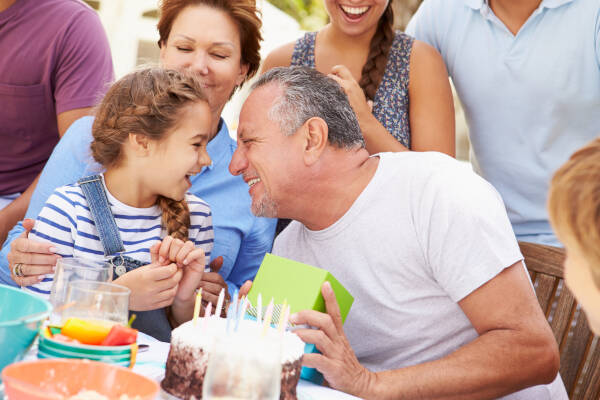 An older man and a young girl sharing a joyful moment at a birthday celebration, surrounded by family members. The table is set with a birthday cake and party decorations.